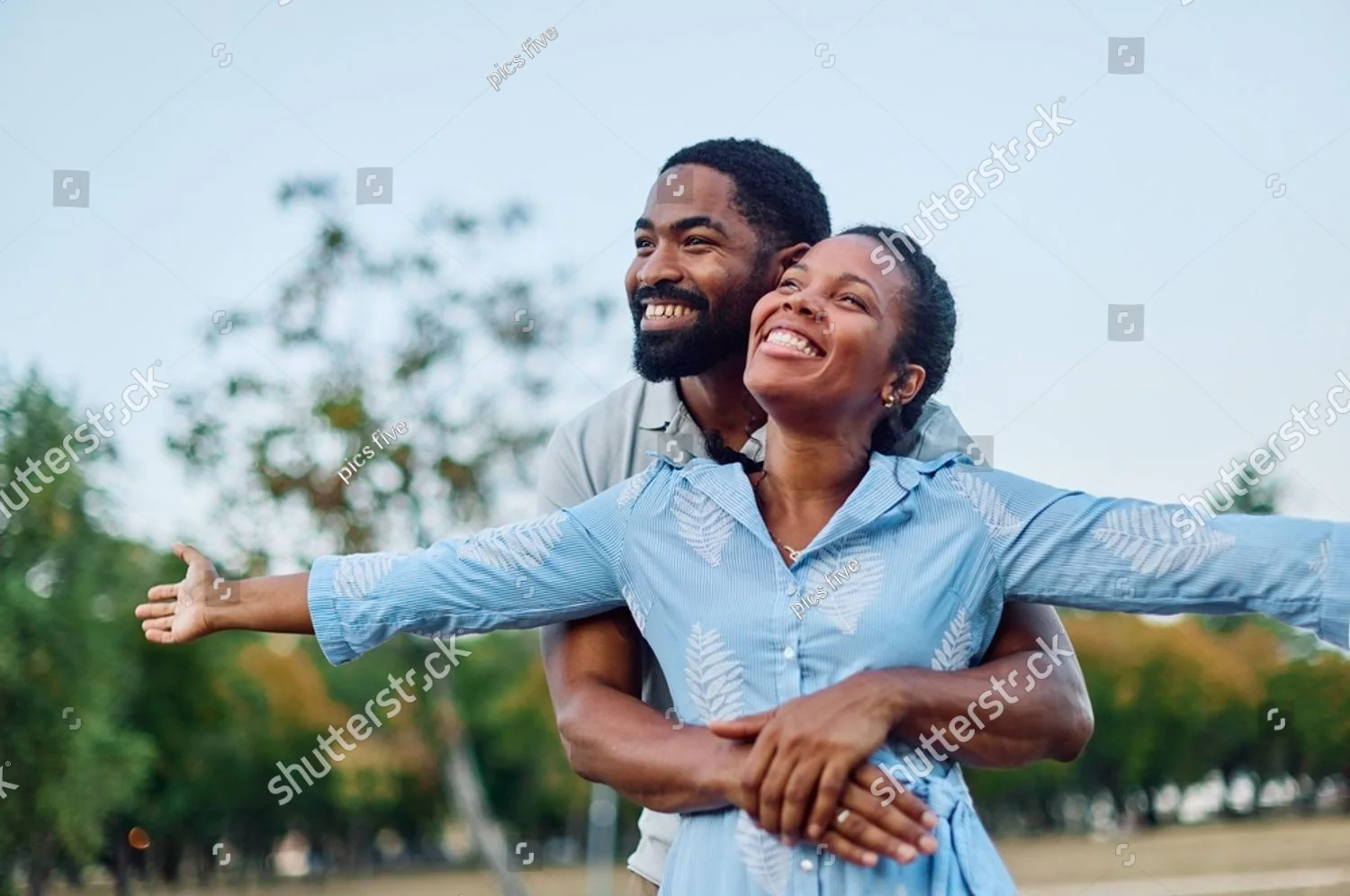 A smiling couple outdoors, his arms around her as she stretches her arms wide